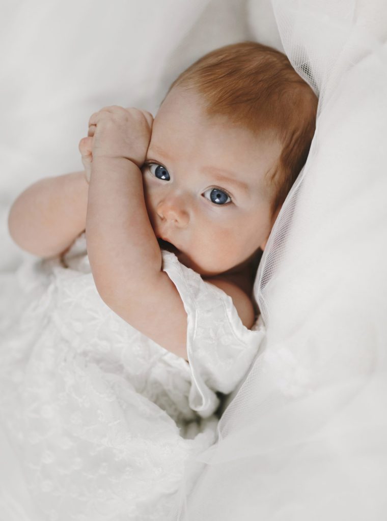 Redhead baby girl is lying on the white blanket with big clear blue eyes, and covering face with tiny arms, happy maternity